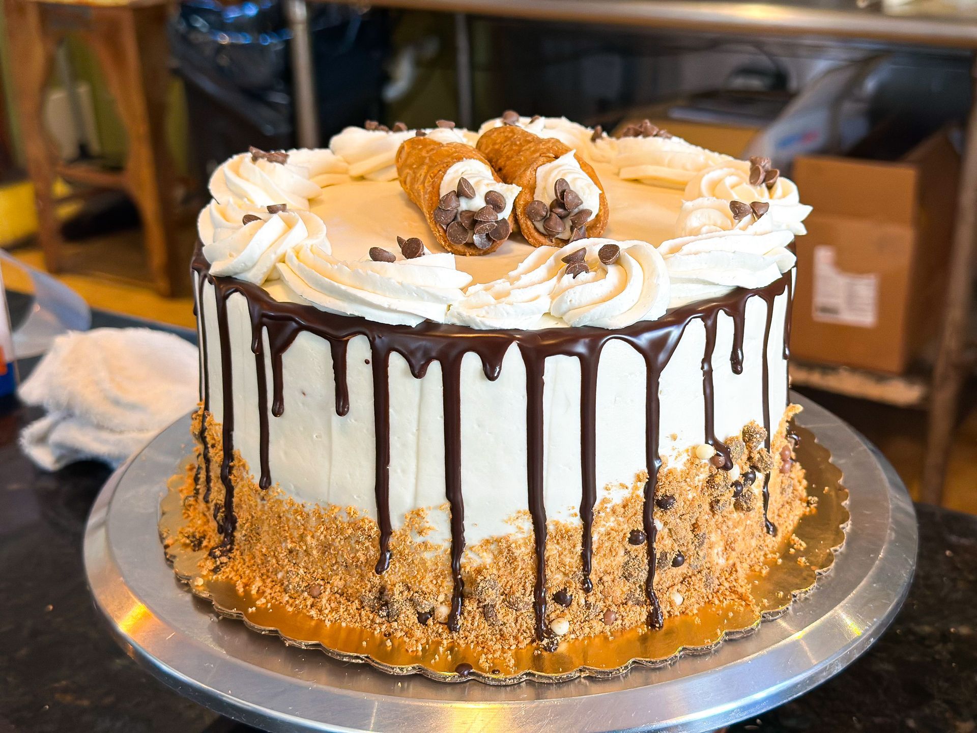 Cake with white frosting, chocolate drips, and cannoli decorations on a silver cake stand.