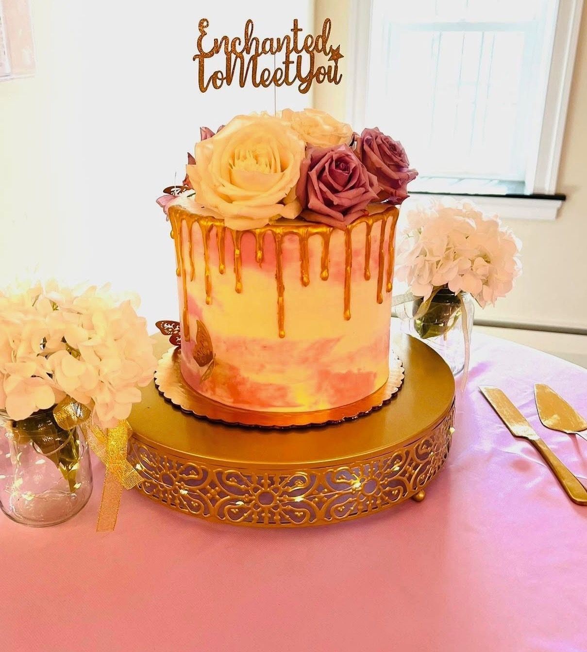 Cake with floral topper, gold drip, on a gold stand, surrounded by flowers, on a pink tablecloth.