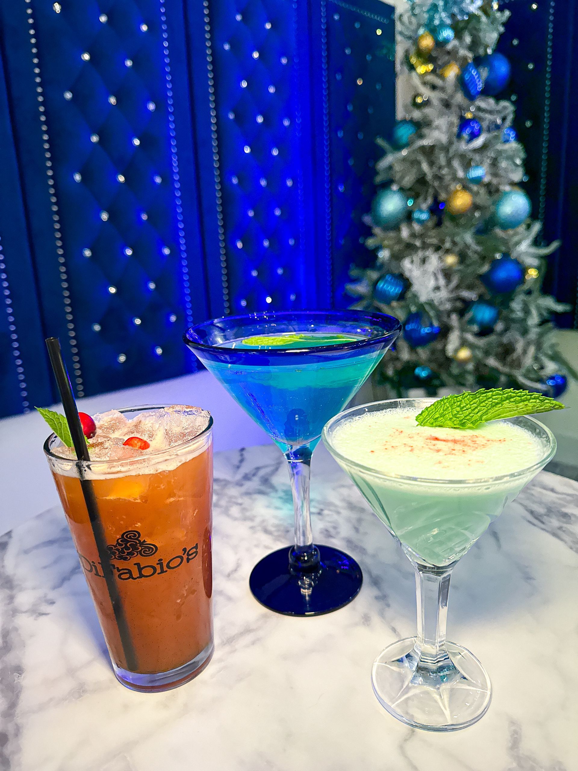 Three colorful cocktails on a marble surface with a blue backdrop and Christmas tree.