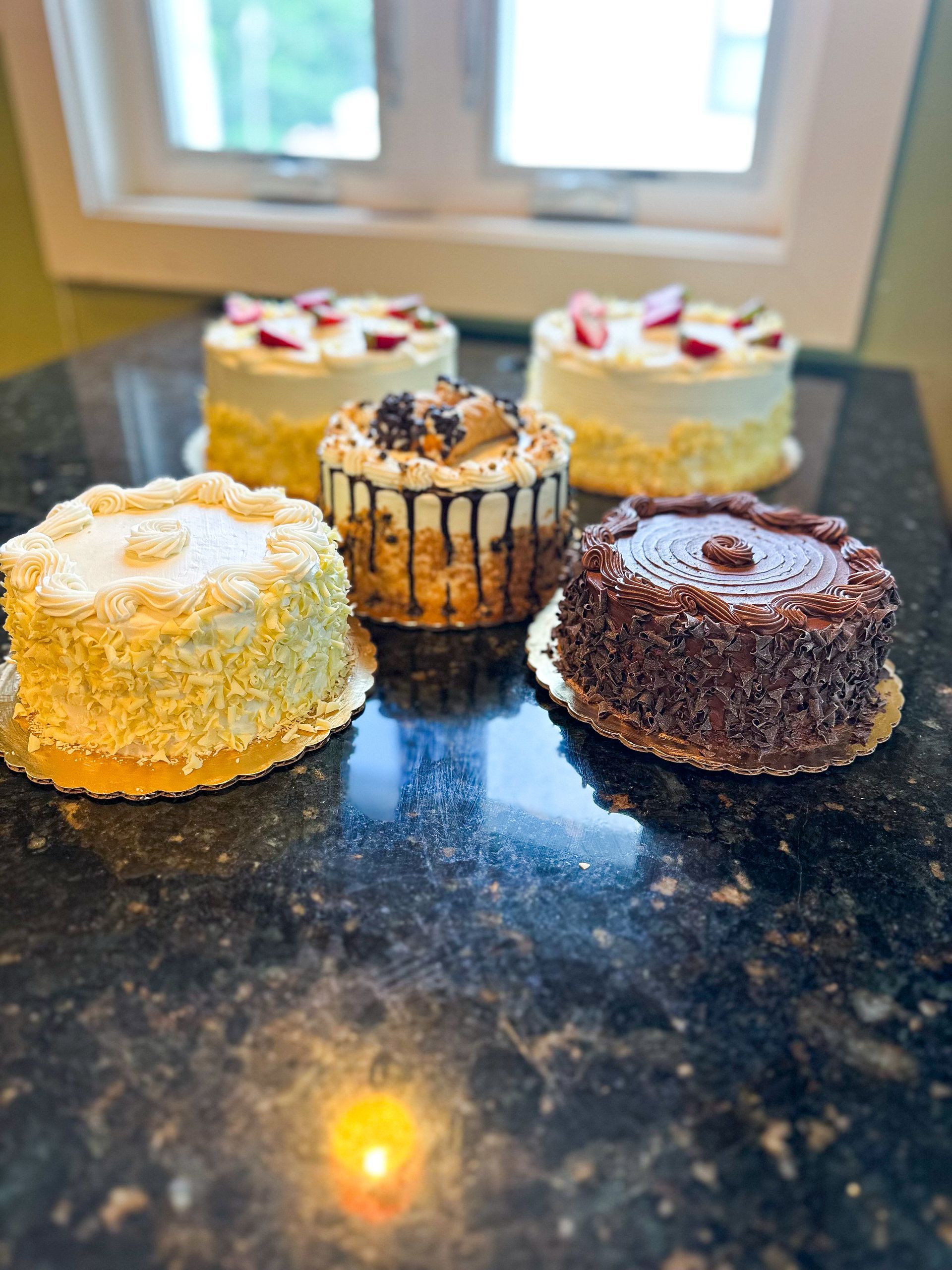 Five small cakes on a countertop; some with strawberries, chocolate, and toppings; near a window.