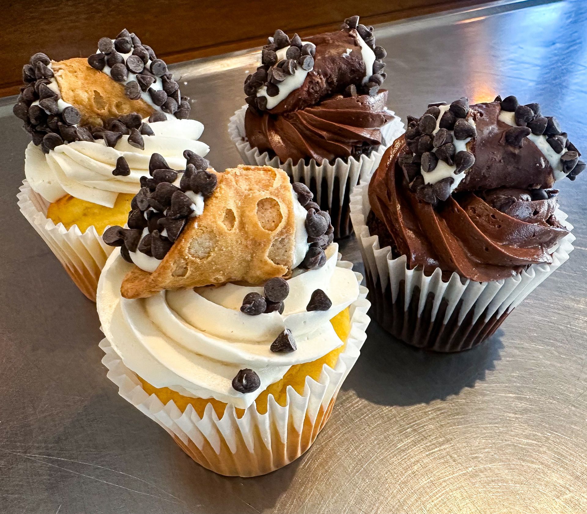 Cupcakes, some topped with chocolate frosting and others with vanilla, garnished with chocolate chips and cannoli shells.