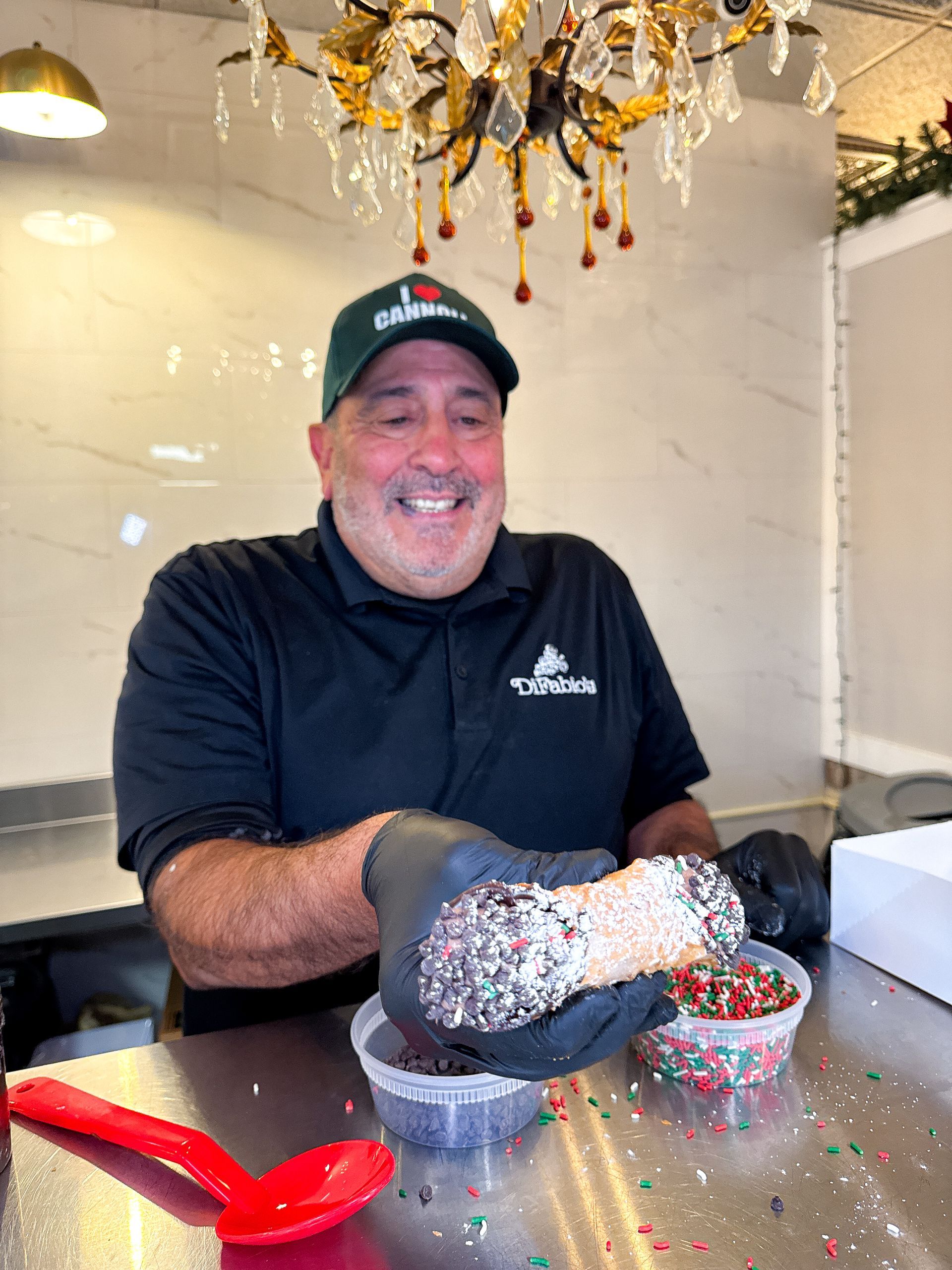 Man smiling, holding a decorated cannoli. He's wearing a green hat and black gloves, near sprinkles and a red spoon.