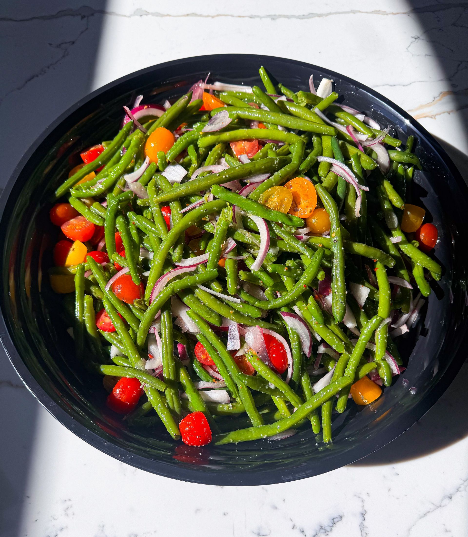 Green bean salad with red and yellow cherry tomatoes and red onion in a black bowl.