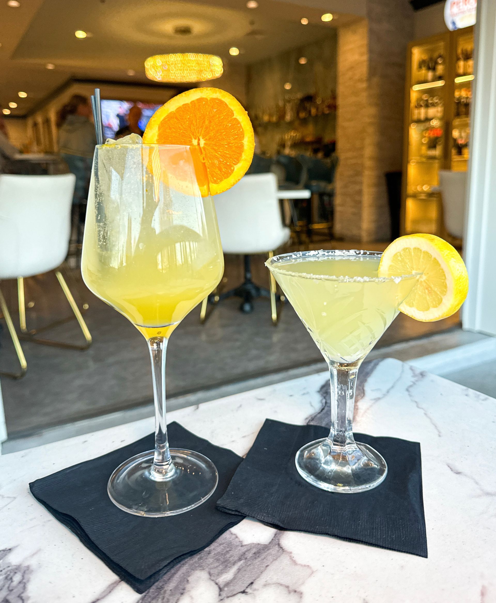 Two cocktails, one in a tall glass with orange, one in a martini glass with lemon, on a bar.