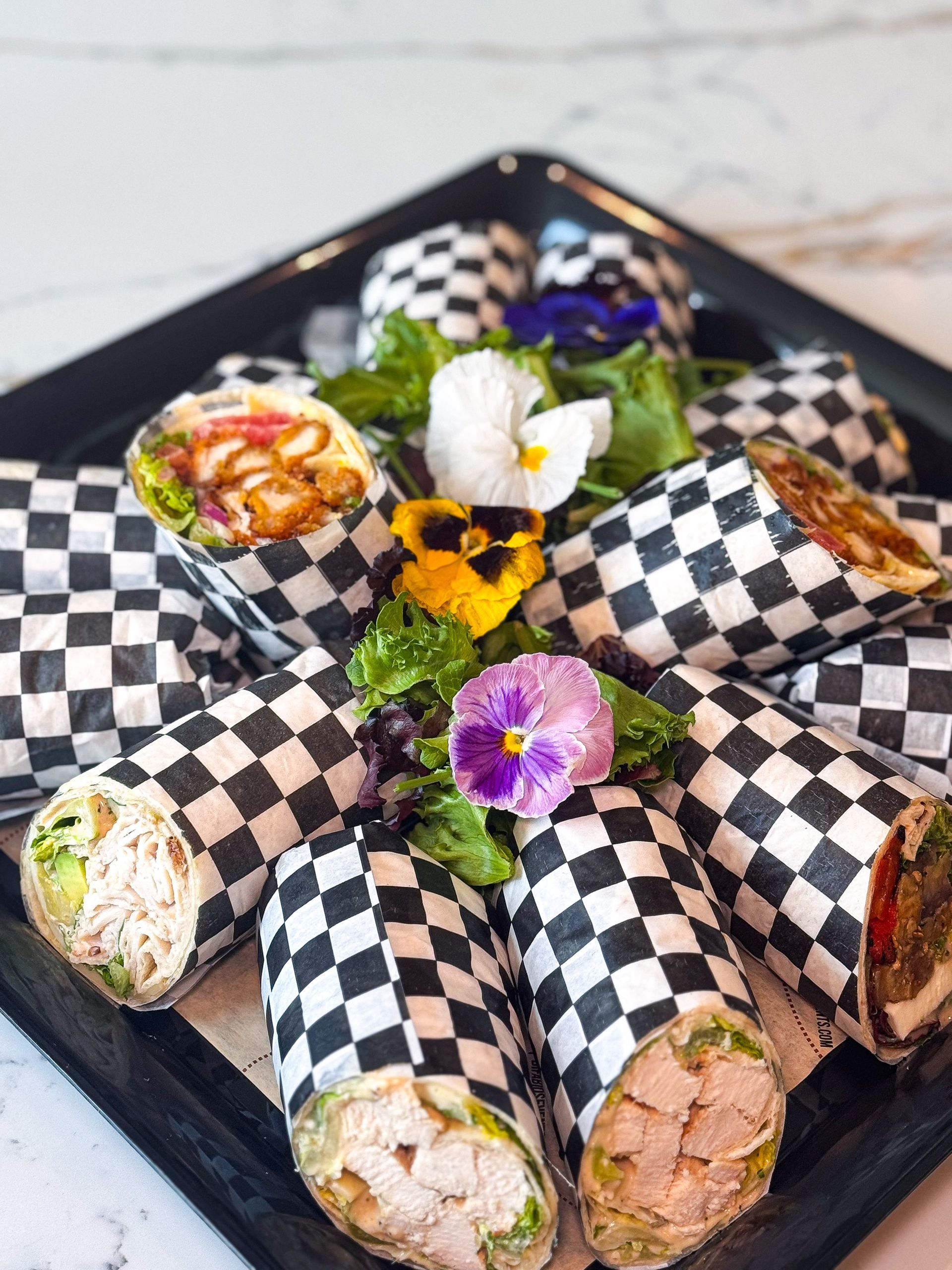 Wraps in checkered paper on a black tray, garnished with flowers and greens.