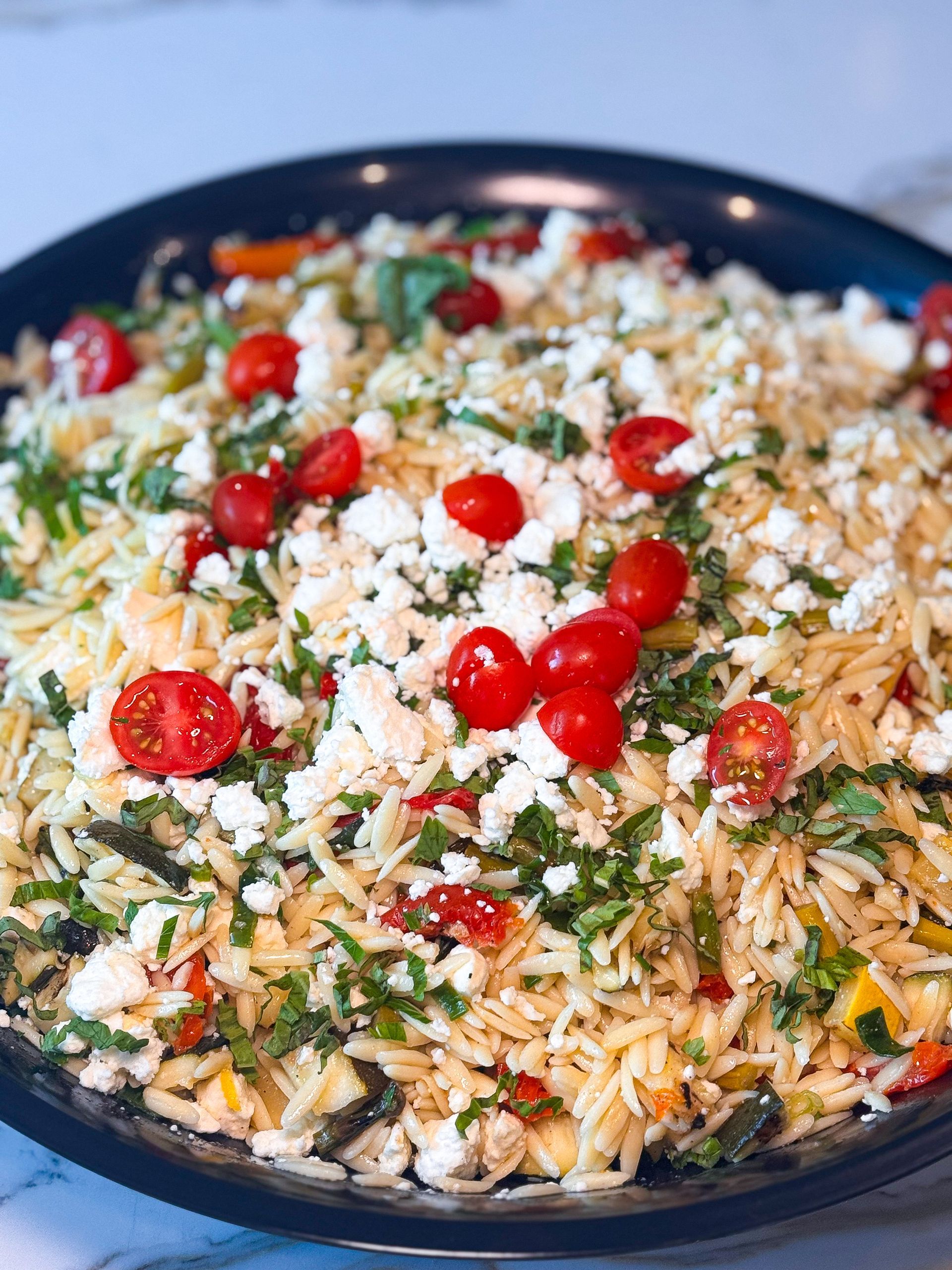 Orzo pasta salad with cherry tomatoes, feta, and basil in a dark blue bowl.