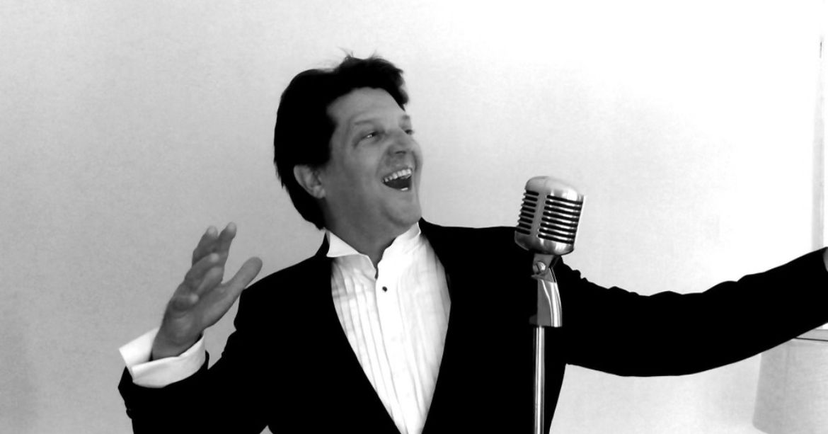 Man in tuxedo singing with vintage microphone, arms outstretched, smiling.
