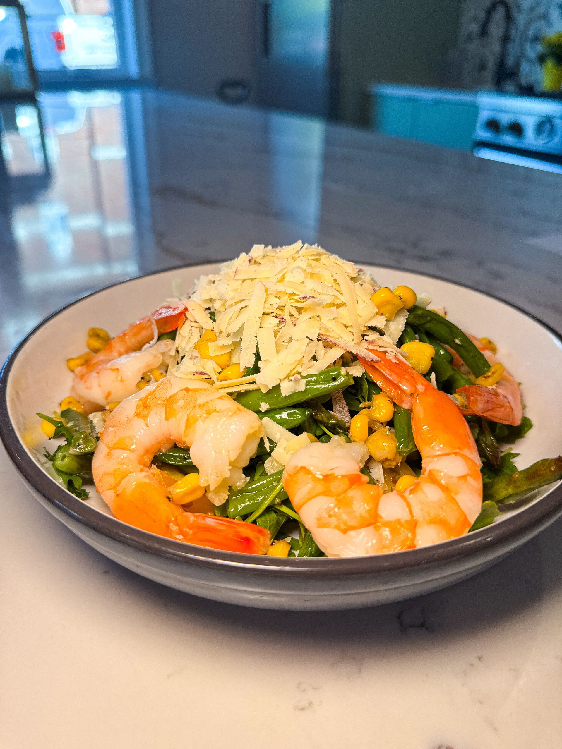 Shrimp, green beans, and corn salad topped with shredded cheese in a gray bowl on a marble countertop.