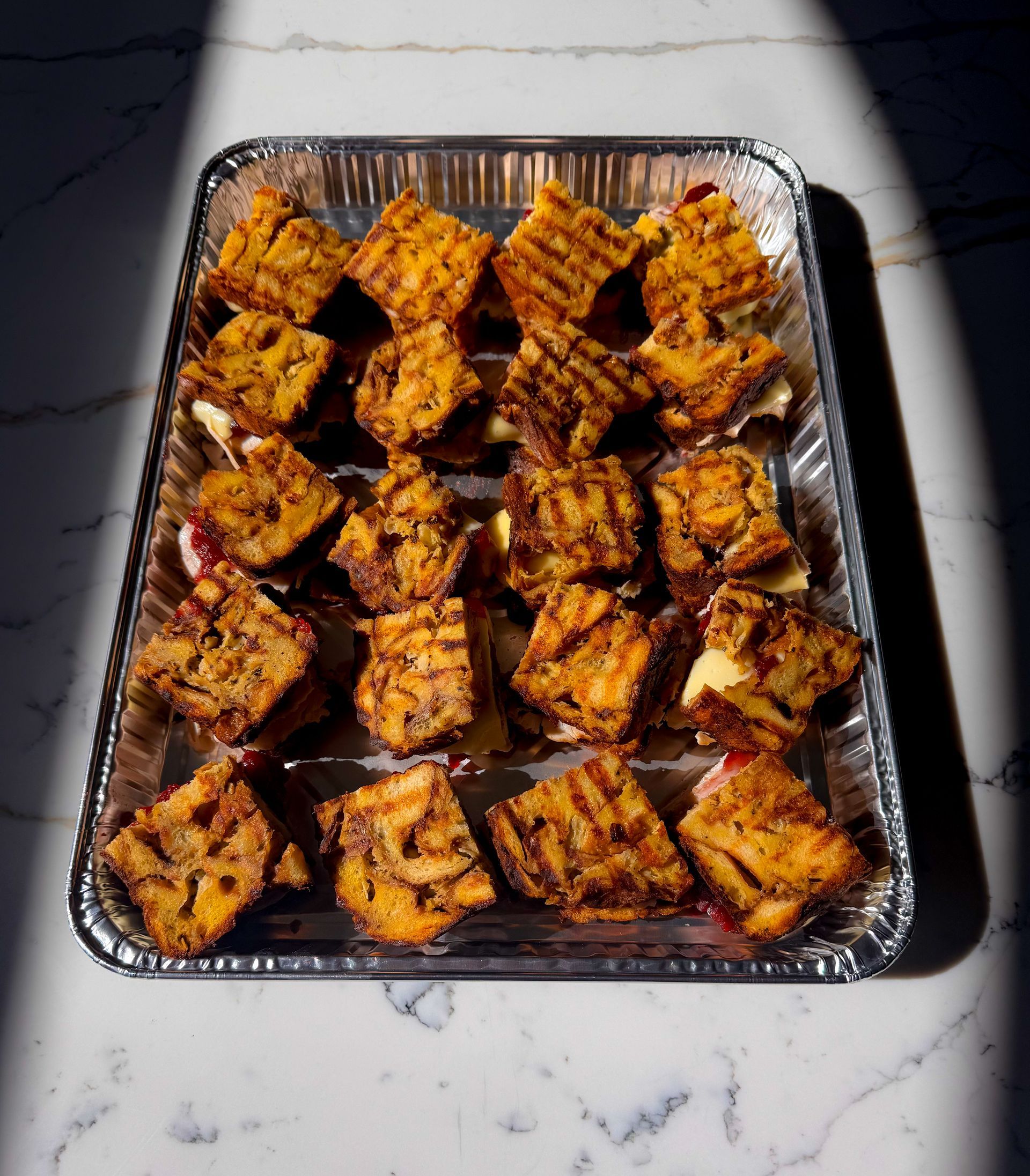 Tray of waffle sandwiches with grilled tops, in a foil container, lit by angled sunlight.