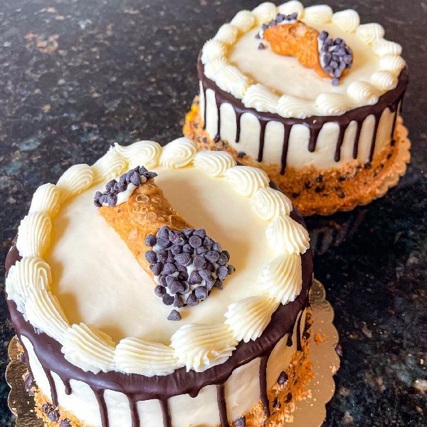 Two round cakes topped with cream, chocolate drip, a cannoli, and chocolate chips.
