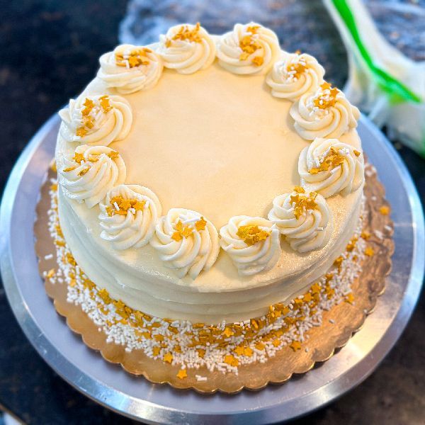 Round frosted cake with cream-colored icing, decorated with piped swirls and yellow sprinkles, on a gold cake board.