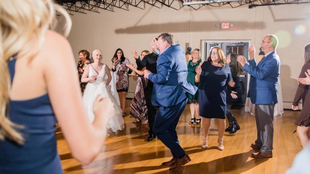 People dancing on a wooden floor at a wedding reception; some are clapping, others smiling.