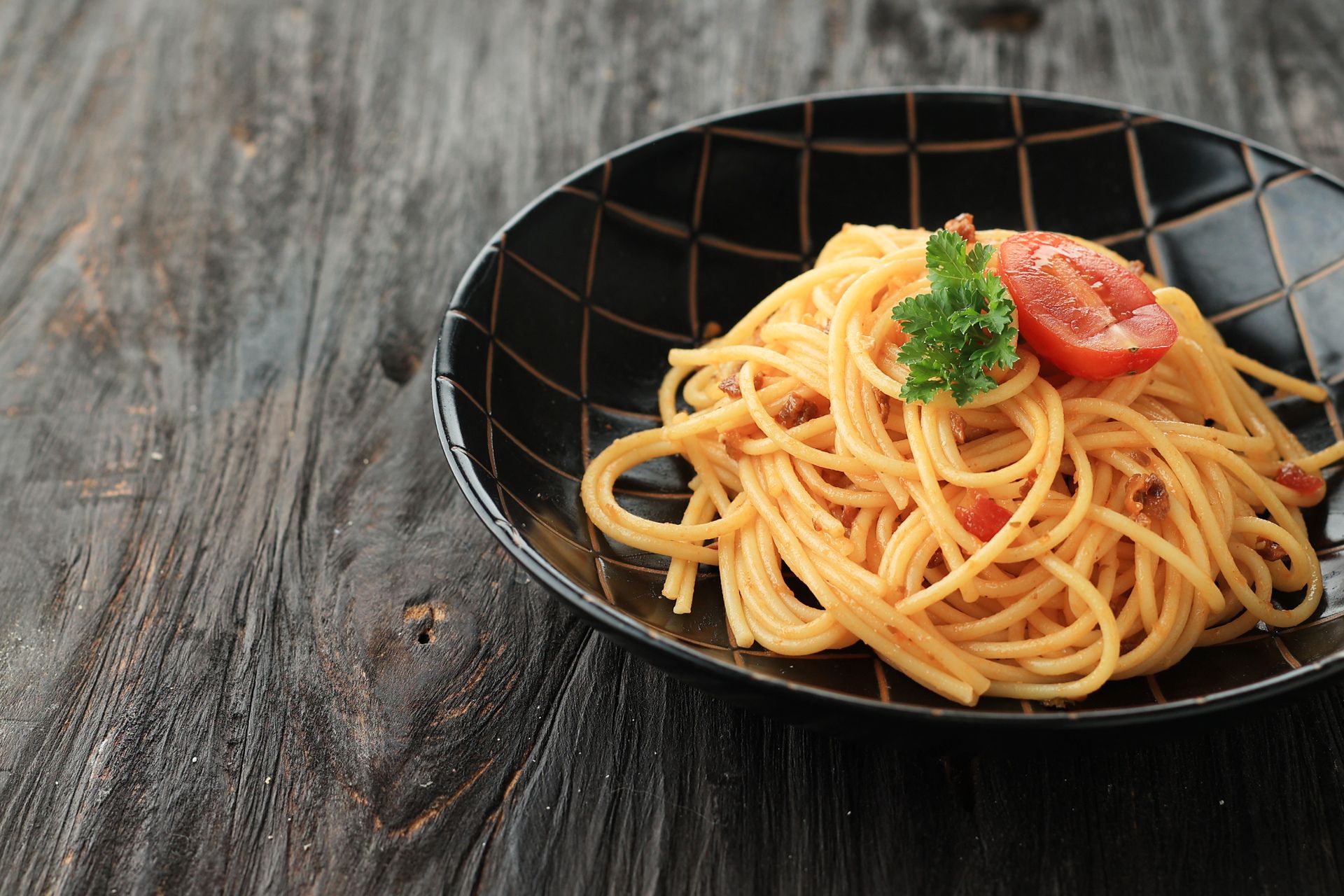 Spaghetti pasta in a black bowl, garnished with tomato and parsley, on a dark wood table.