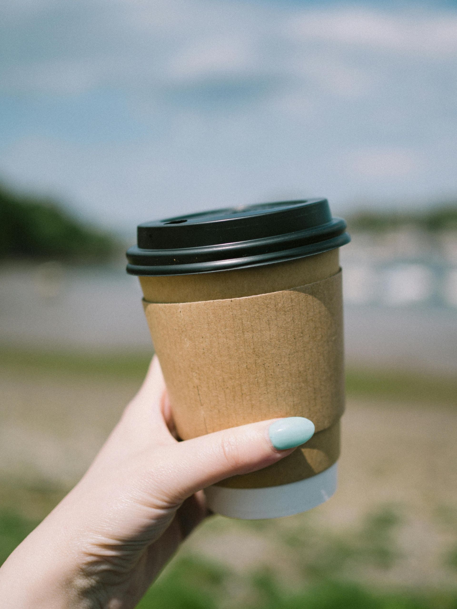Hand holding a takeaway coffee cup with a brown sleeve, outdoors on a sunny day.