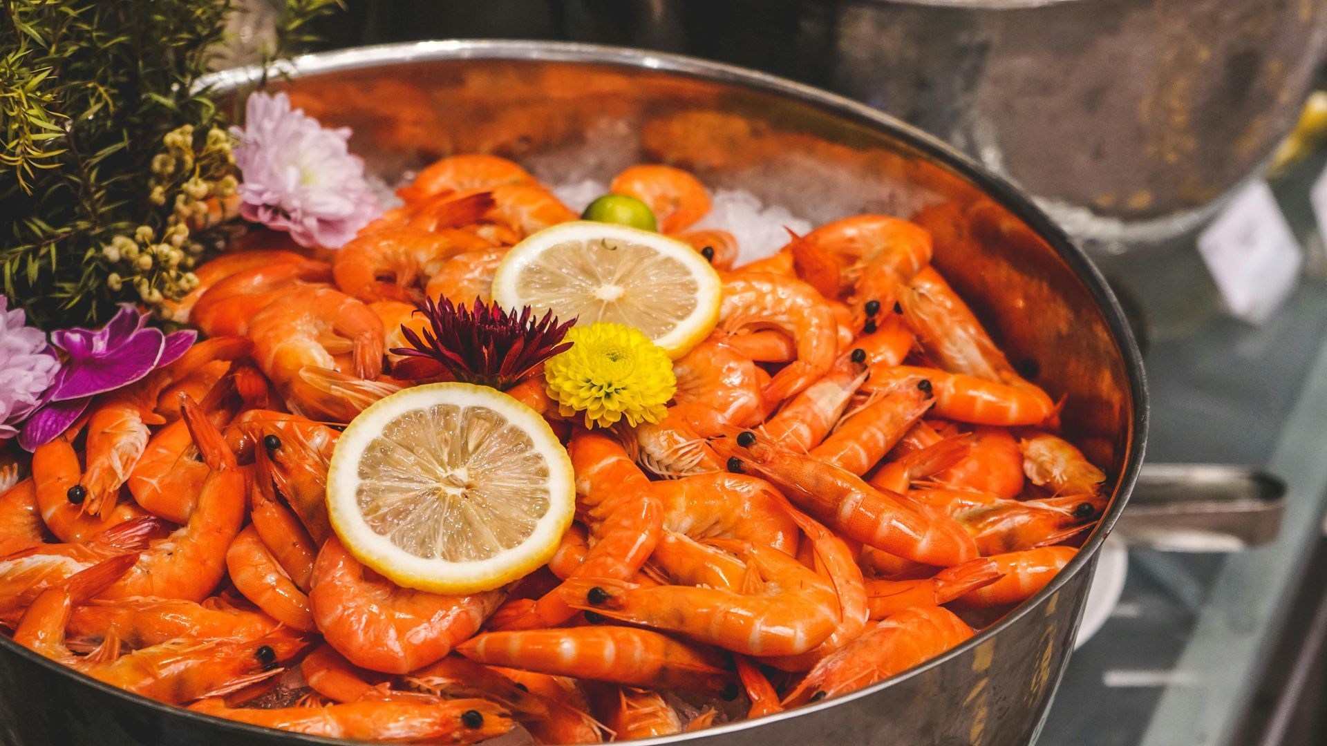Cooked shrimp in a metal bowl, garnished with lemon slices and flowers.