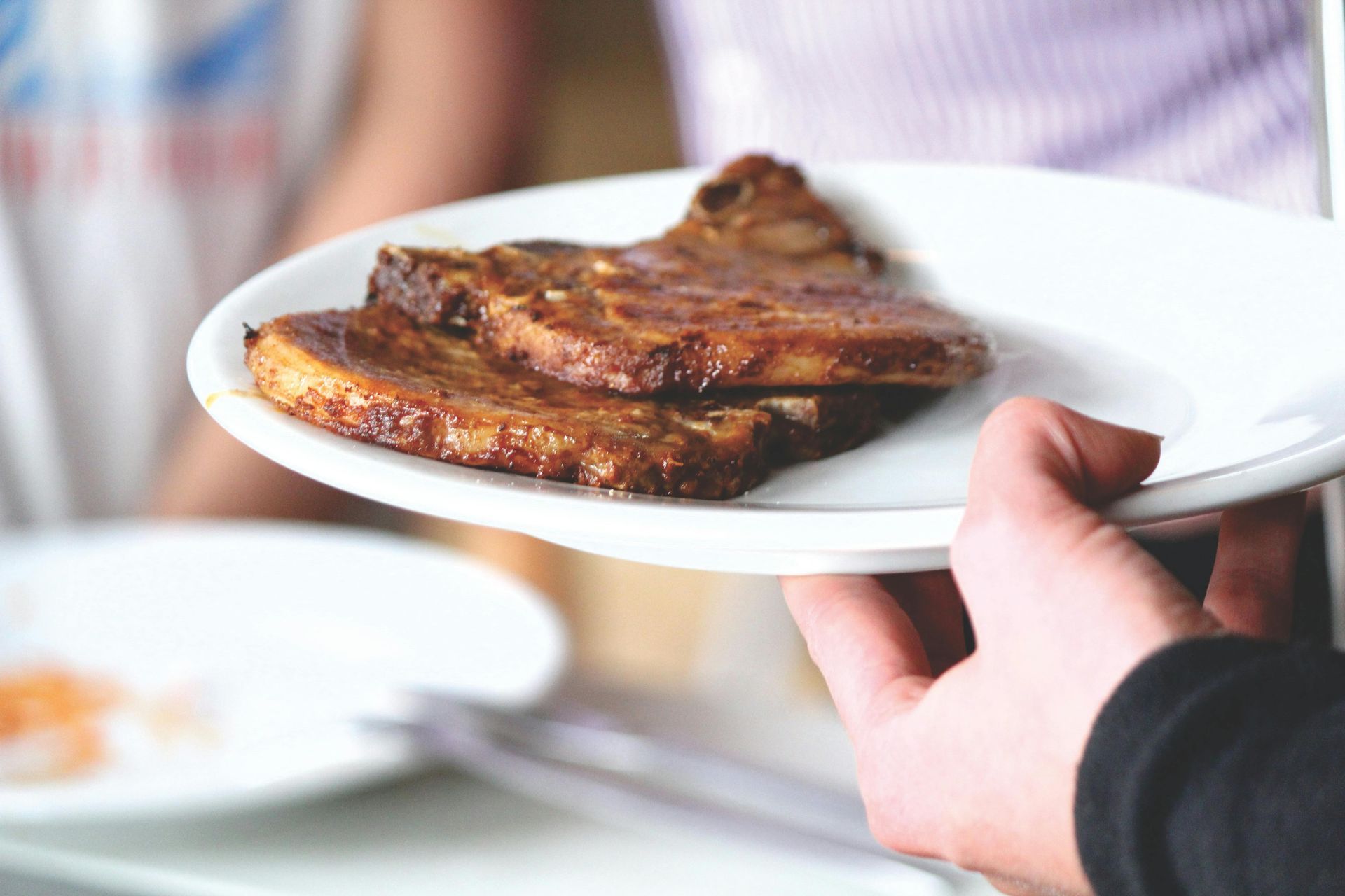 Hand holding a plate with two cooked pork chops. Another plate and person in background.