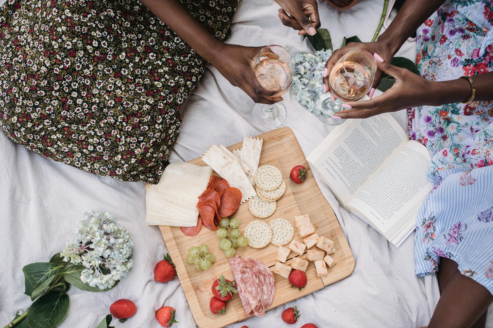 Picnic with charcuterie board, drinks, and book. Two people hold glasses, surrounded by food, flowers, and blanket.