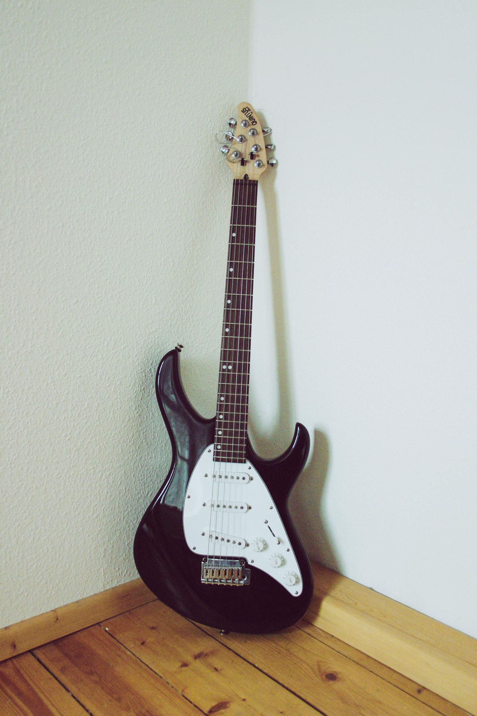 Black electric guitar leaning in a corner against white walls and wood floor.