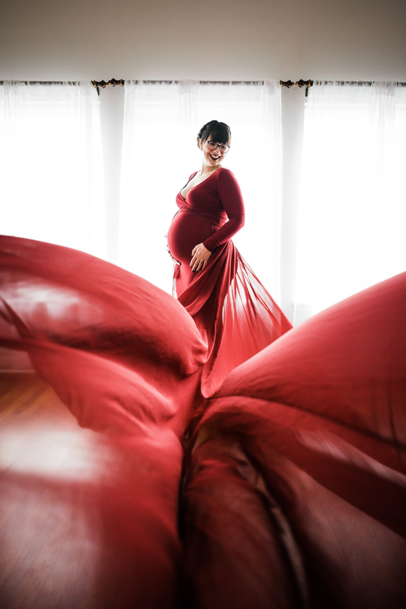 A pregnant woman in a long red dress is standing in front of a window.