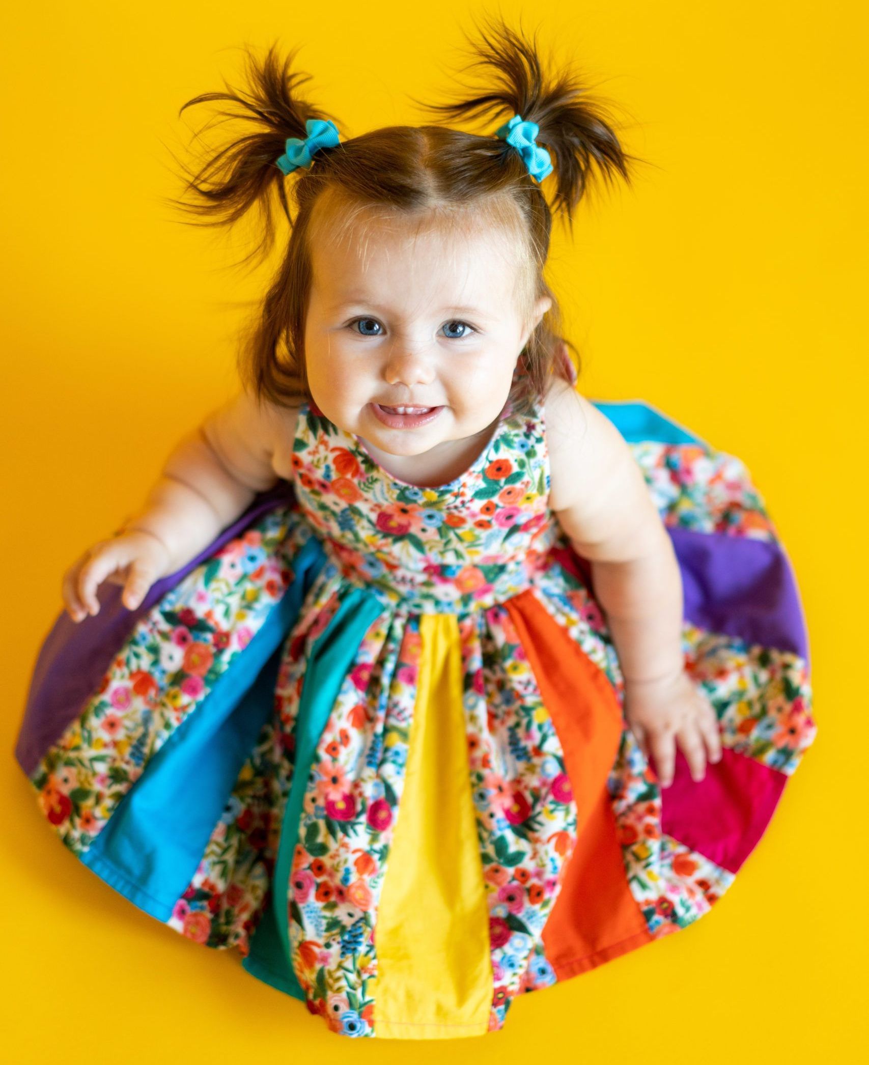 A baby girl wearing a colorful dress is sitting on a yellow surface.