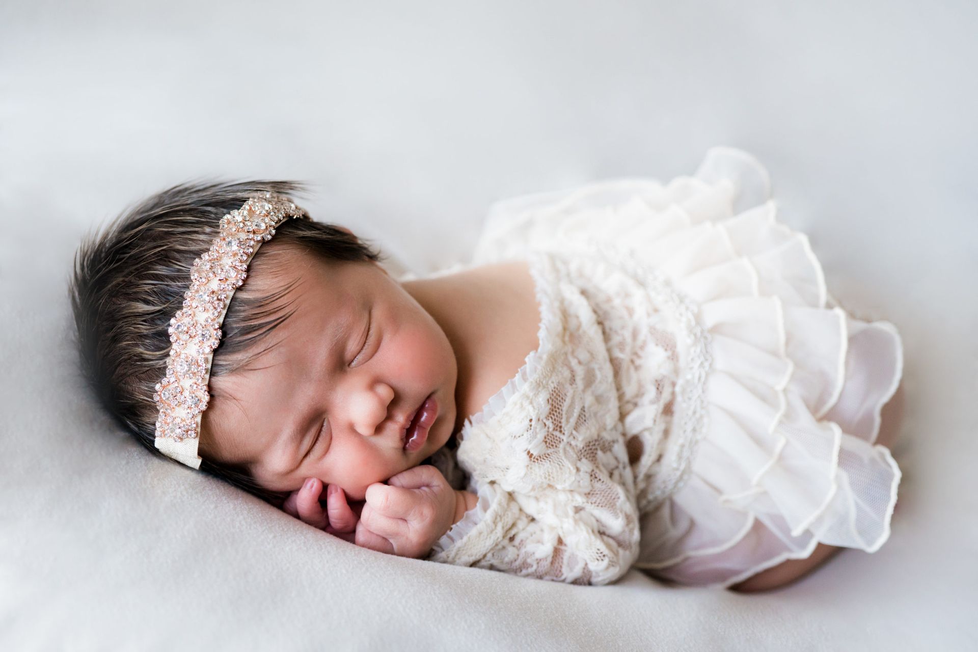 A newborn baby girl wearing a white dress and headband is sleeping on a bed.