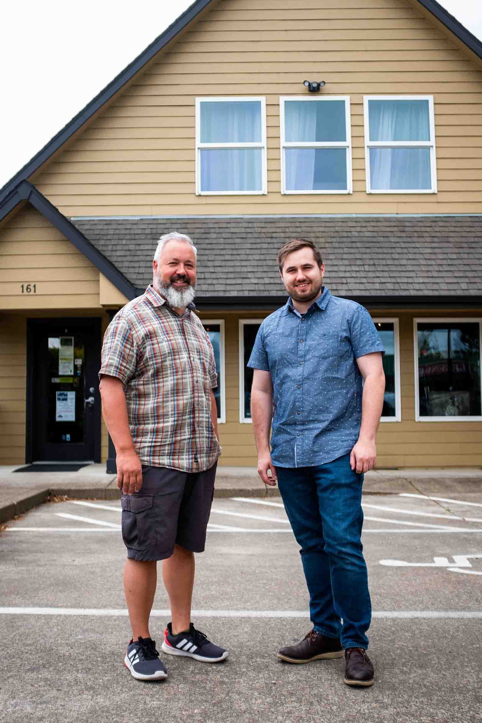 Two men are standing in front of a building in a parking lot.