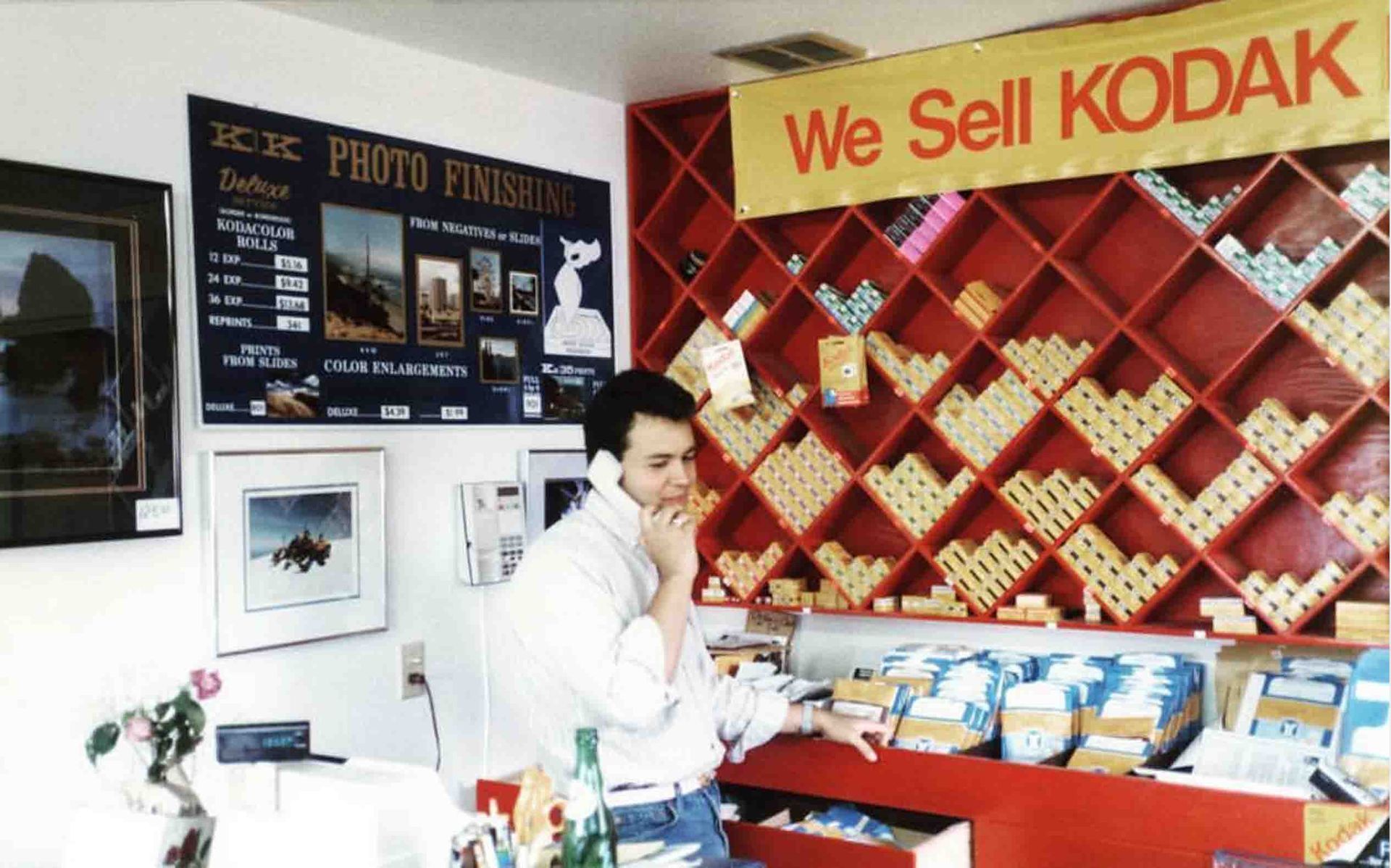 A man talking on a cell phone in front of a sign that says we sell kodak