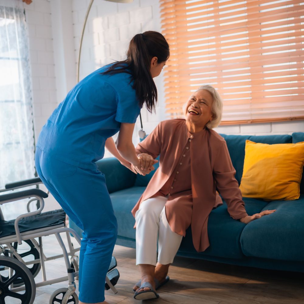 A nurse is helping an elderly woman in a wheelchair sit on a couch.