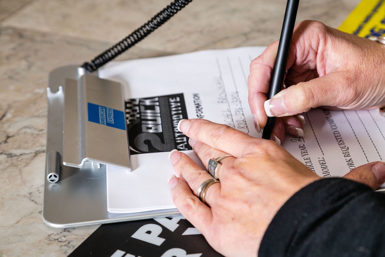 A close-up of a person writing on a paper attached to a silver clipboard on a marble surface. | 2 QuickAuto, Car & Tire Repair | Copperfield