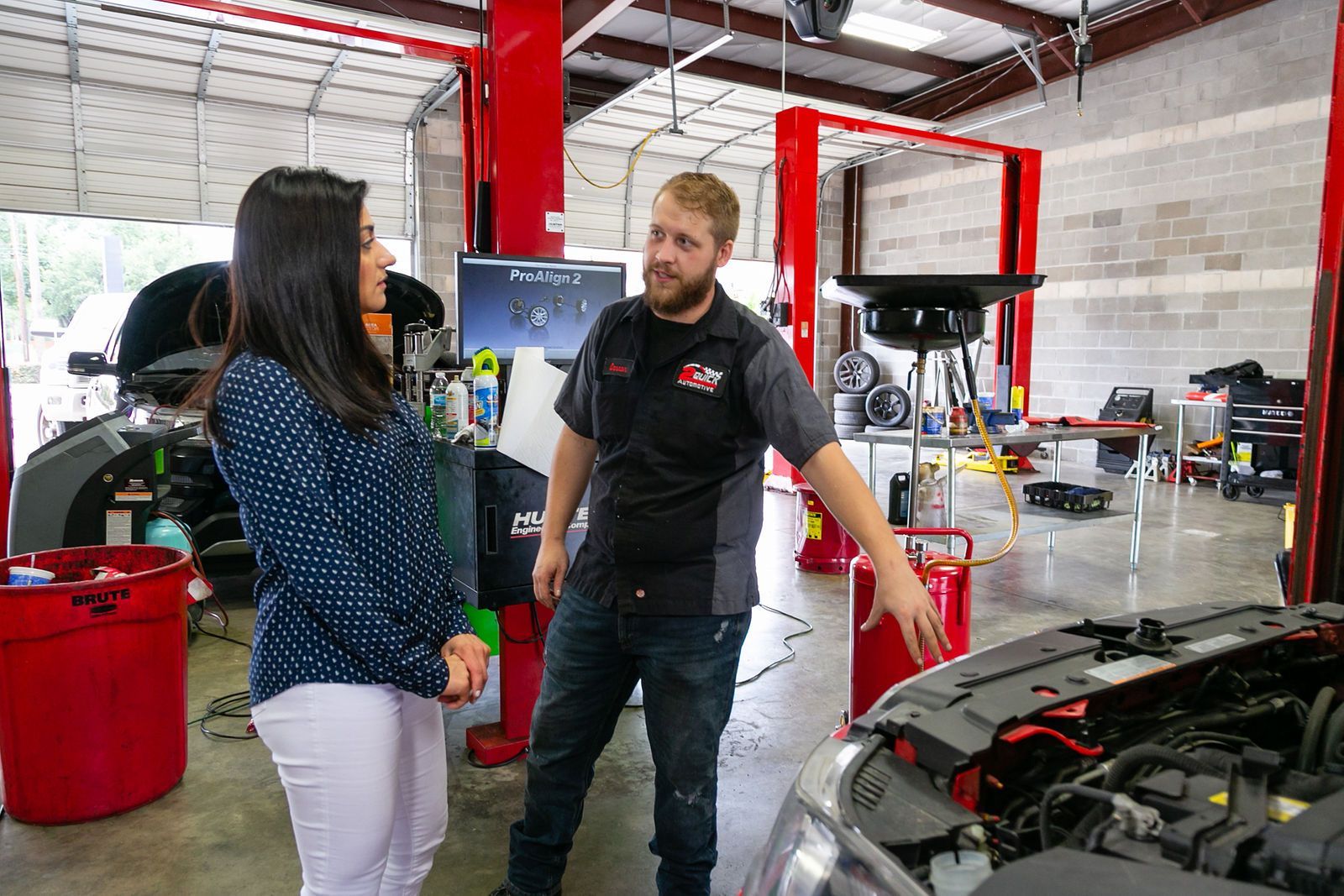 A woman and a mechanic stand in an auto repair shop; the mechanic gestures toward an open car hood. | 2 QuickAuto, Car & Tire Repair | Copperfield