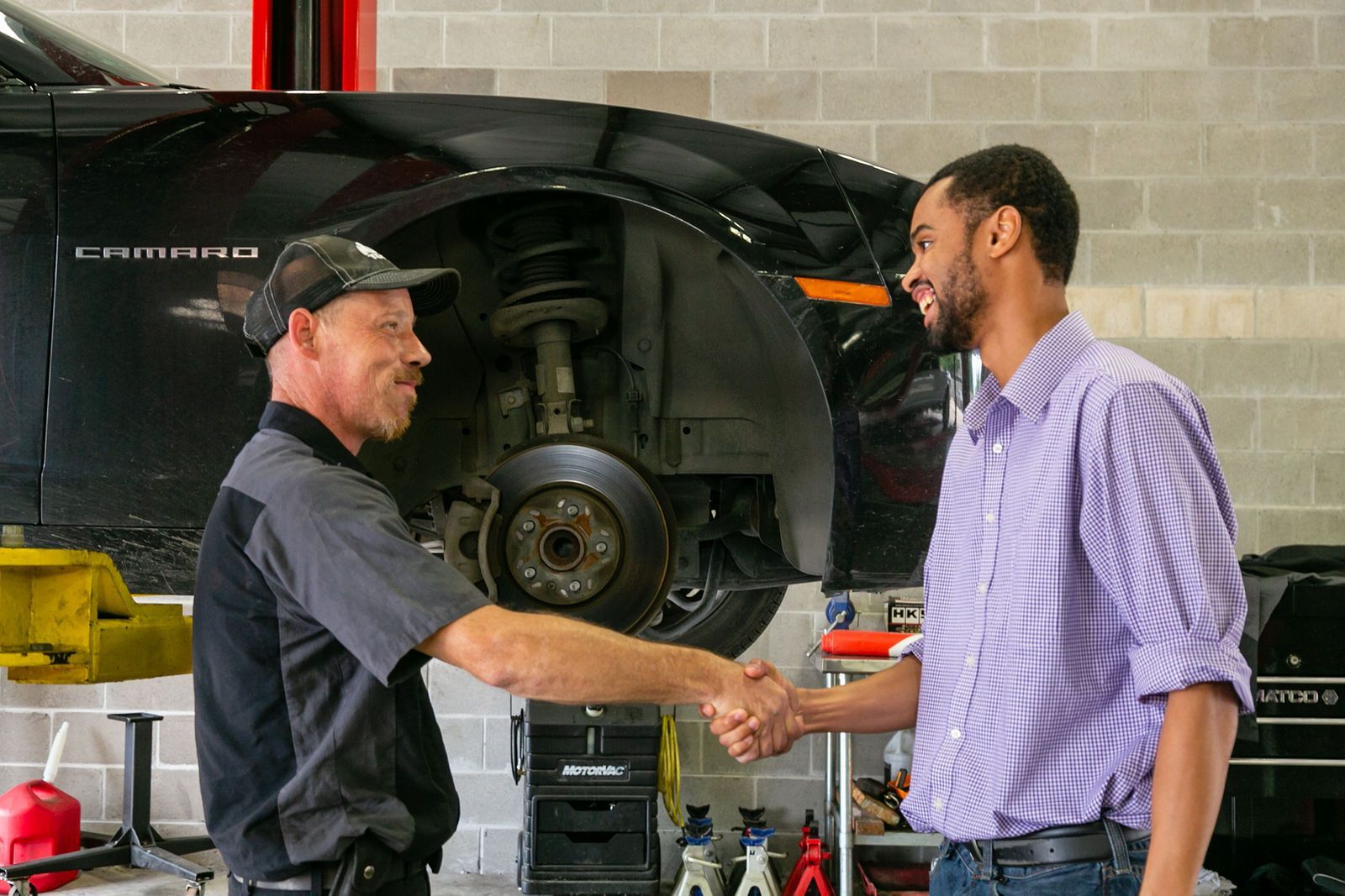 A mechanic in a grey uniform and a customer in a purple checkered shirt shake hands in an auto repair shop. | 2 QuickAuto, Car & Tire Repair | Copperfield