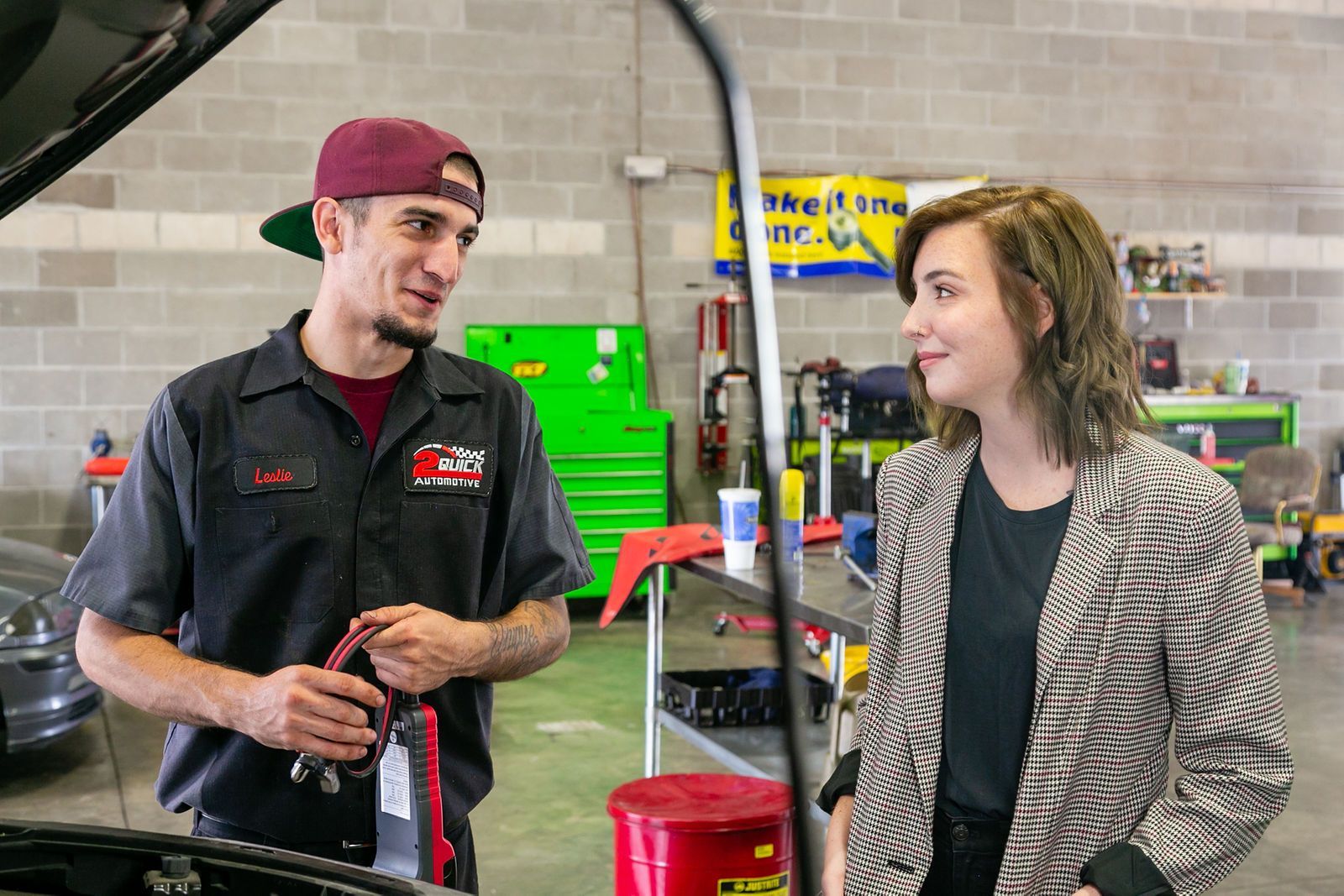 An auto mechanic in a black uniform and maroon cap talks to a person in a patterned blazer inside a garage. | 2 QuickAuto, Car & Tire Repair | Copperfield