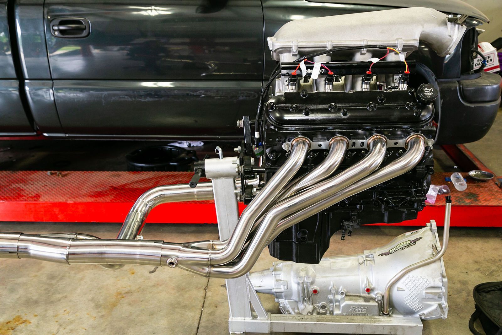 A black internal combustion engine with shiny exhaust headers sits on a stand in a shop with a vehicle in the background. | 2 QuickAuto, Car & Tire Repair | Copperfield