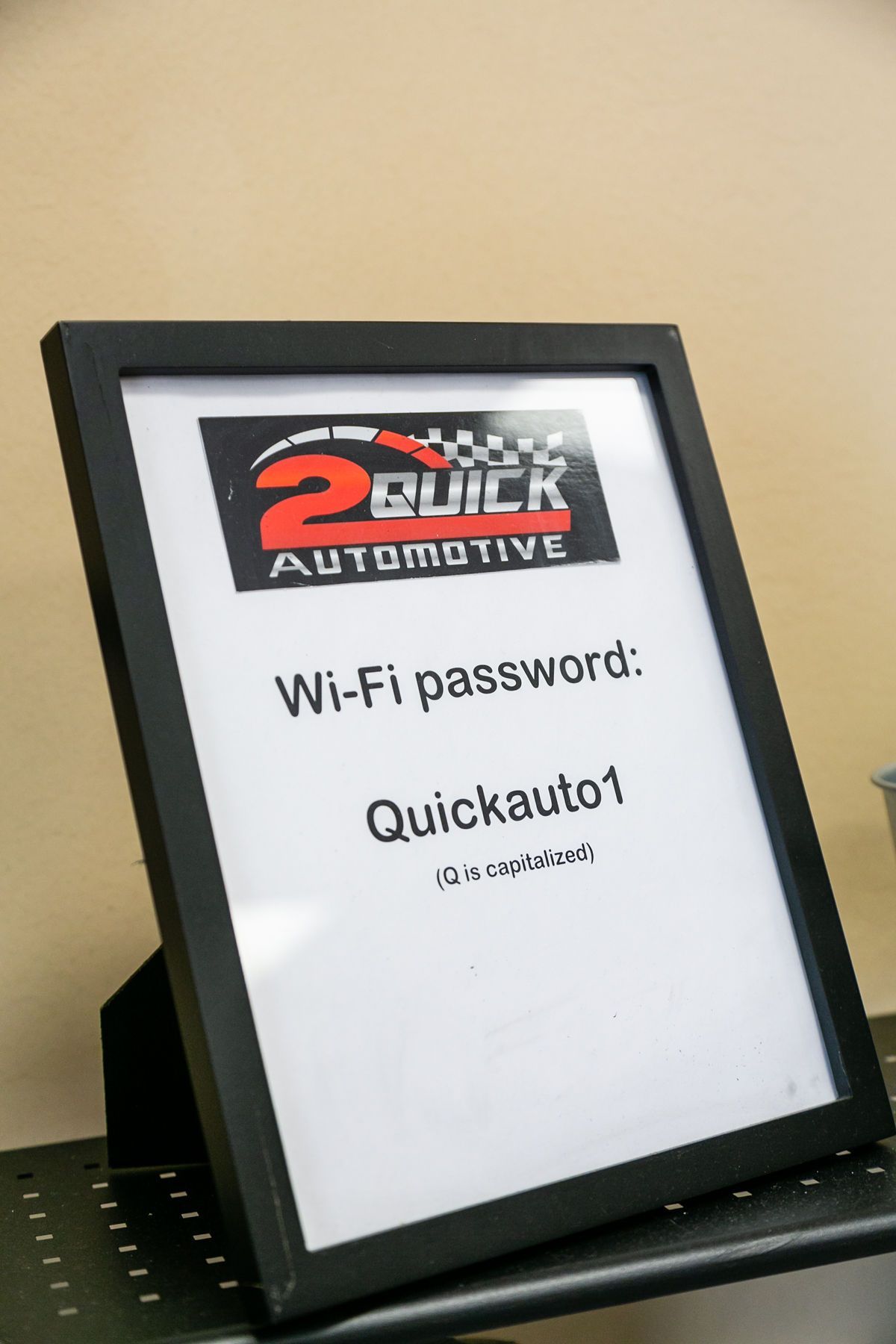 A black-framed sign for 2Quick Automotive on a table, displaying the Wi-Fi password: 