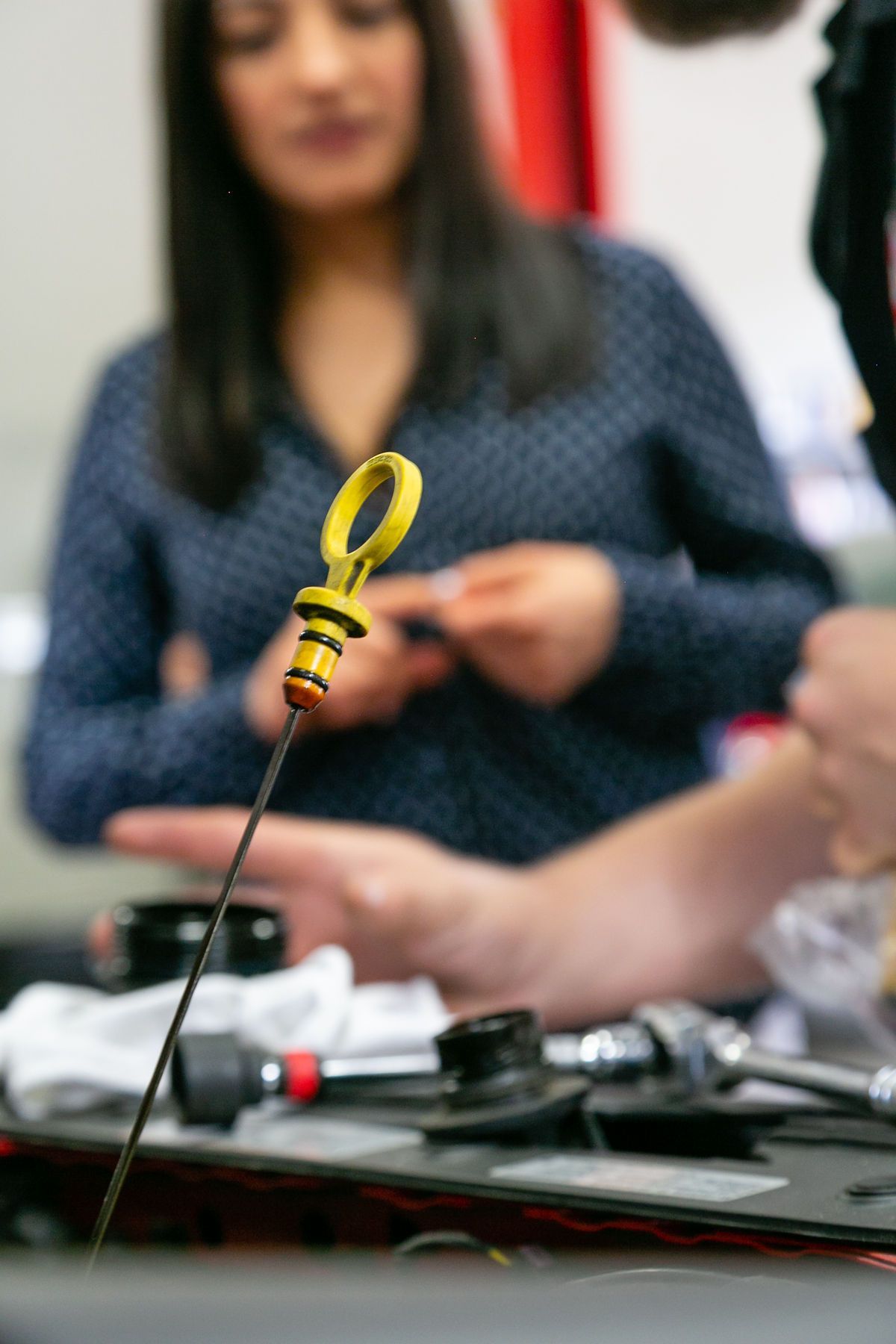 A close-up of a yellow oil dipstick held in the foreground, with blurred people working on an engine in the background. | 2 QuickAuto, Car & Tire Repair | Copperfield