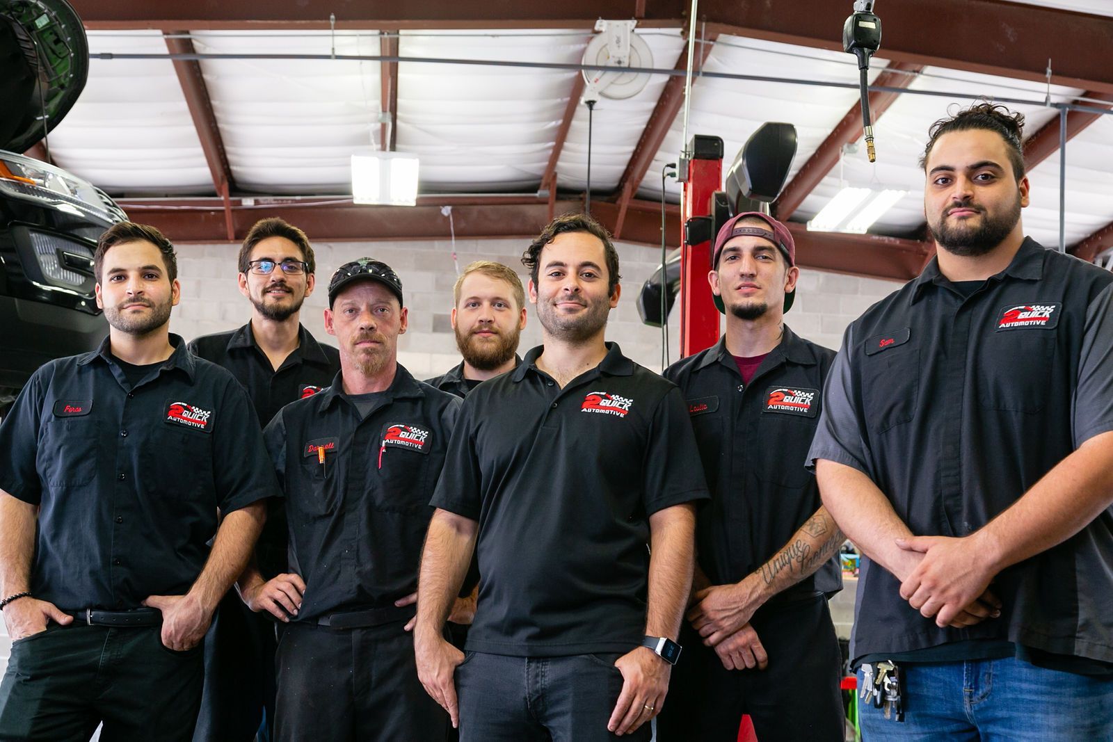 A group of seven auto mechanics wearing black uniform shirts stand together in an automotive repair shop. | 2 QuickAuto, Car & Tire Repair | Copperfield