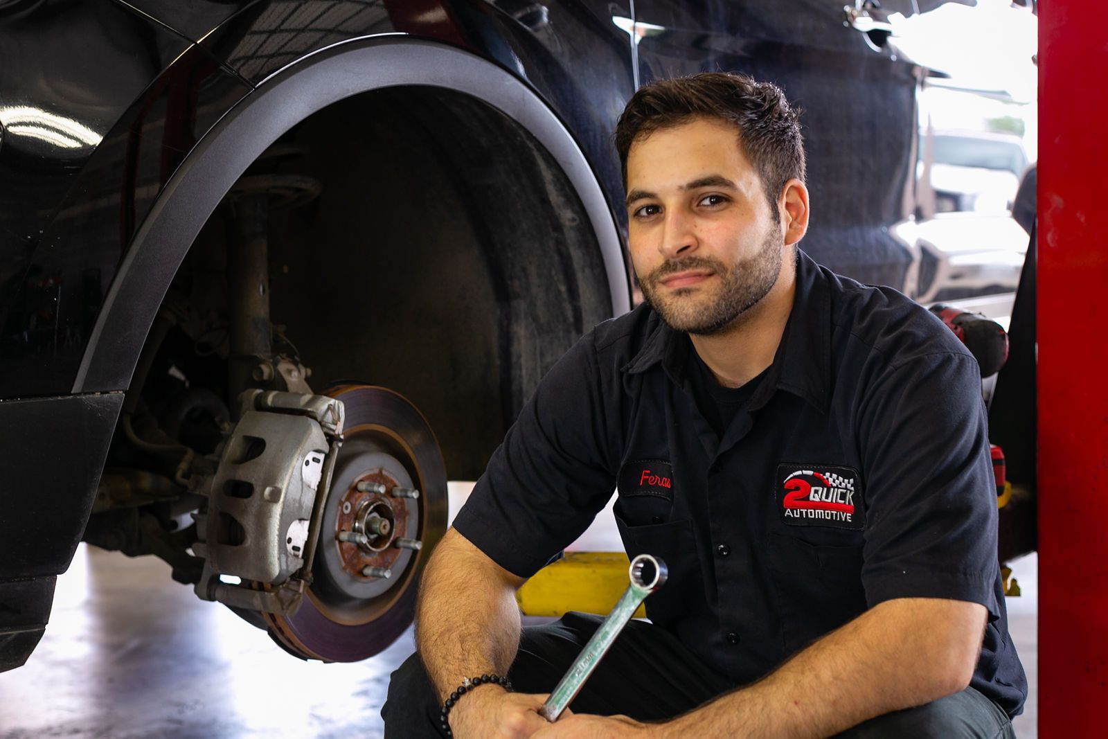 A mechanic wearing a dark uniform holds a wrench while kneeling beside a vehicle with its wheel removed in a garage. | 2 QuickAuto, Car & Tire Repair | Copperfield
