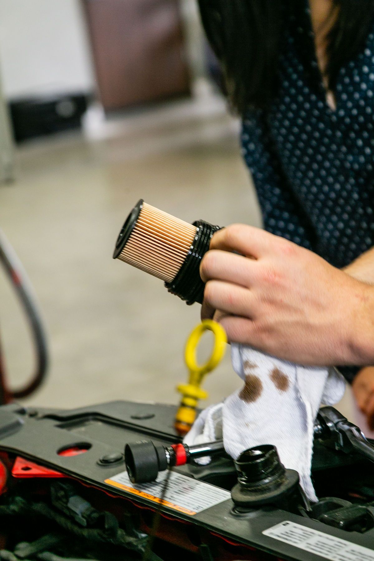 A person holds a cylindrical car oil filter above an engine bay, using a white cloth to wipe an oil dipstick. | 2 QuickAuto, Car & Tire Repair | Copperfield