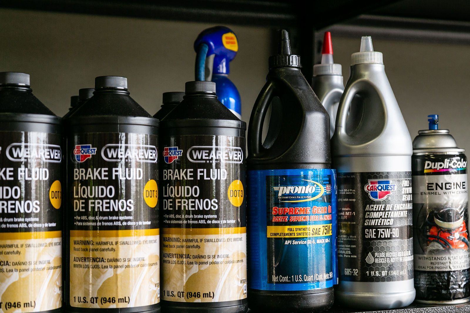 A row of automotive fluid bottles, including brake fluid, gear oil, and an aerosol can, arranged on a garage shelf. | 2 QuickAuto, Car & Tire Repair | Copperfield