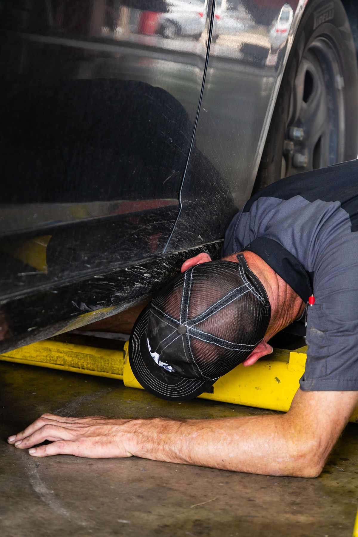 A technician in a uniform and cap kneels under a car on a yellow lift, working in an auto repair shop. | 2 QuickAuto, Car & Tire Repair | Copperfield
