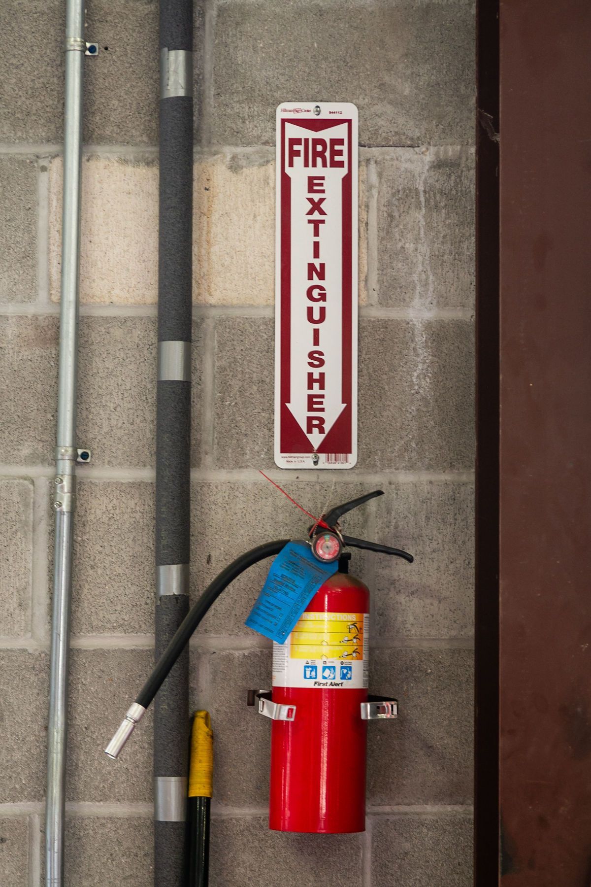 A red fire extinguisher mounted on a grey cinder block wall below a vertical sign pointing downward. | 2 QuickAuto, Car & Tire Repair | Copperfield