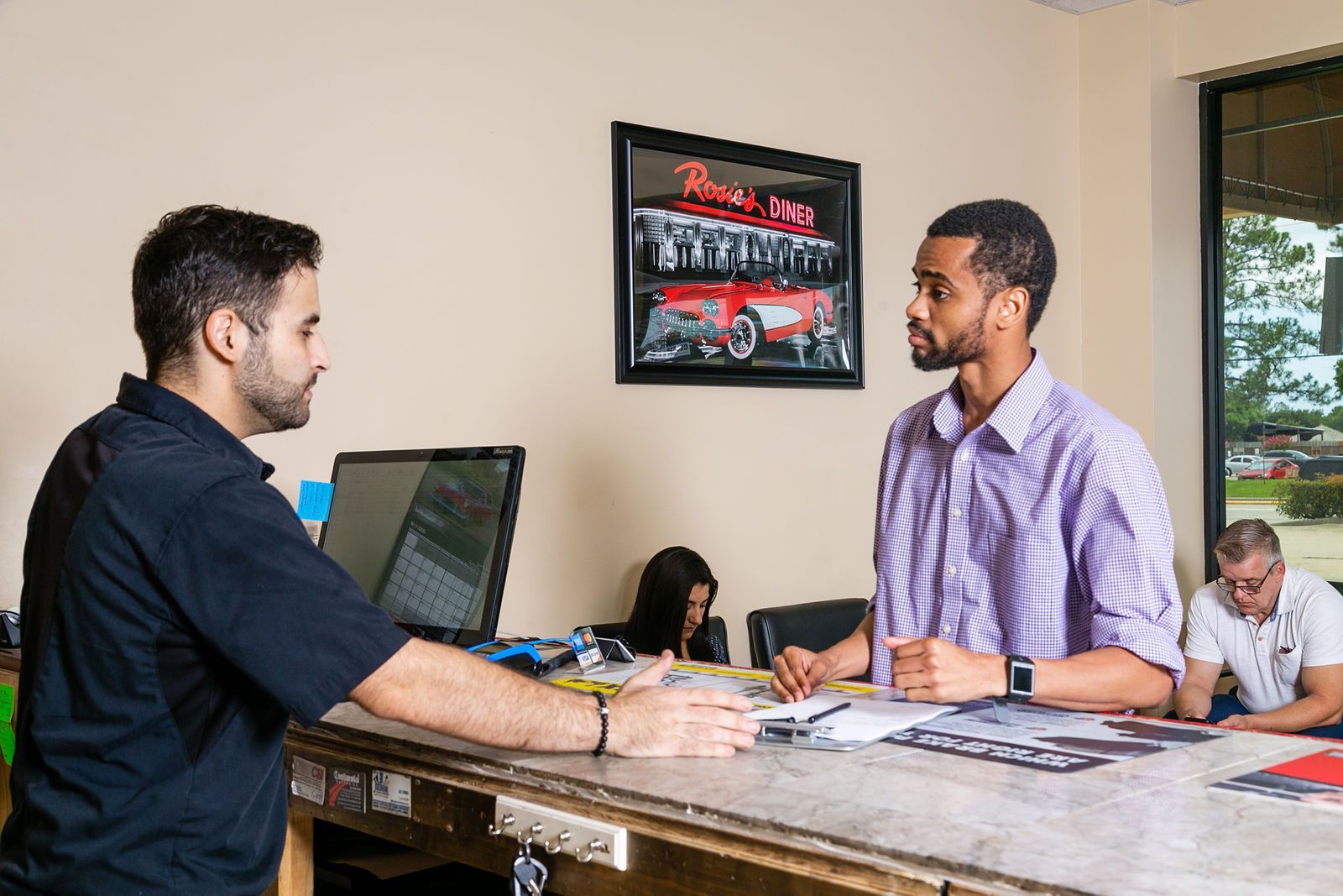 Two people stand at a counter in an office; one is speaking to the other while filling out paperwork. | 2 QuickAuto, Car & Tire Repair | Copperfield