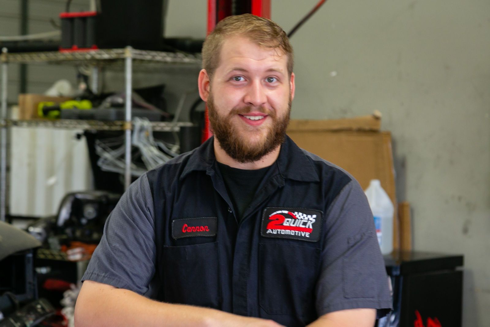 A smiling person with a beard, wearing a gray and black uniform, standing in a workshop setting. | 2 QuickAuto, Car & Tire Repair | Copperfield