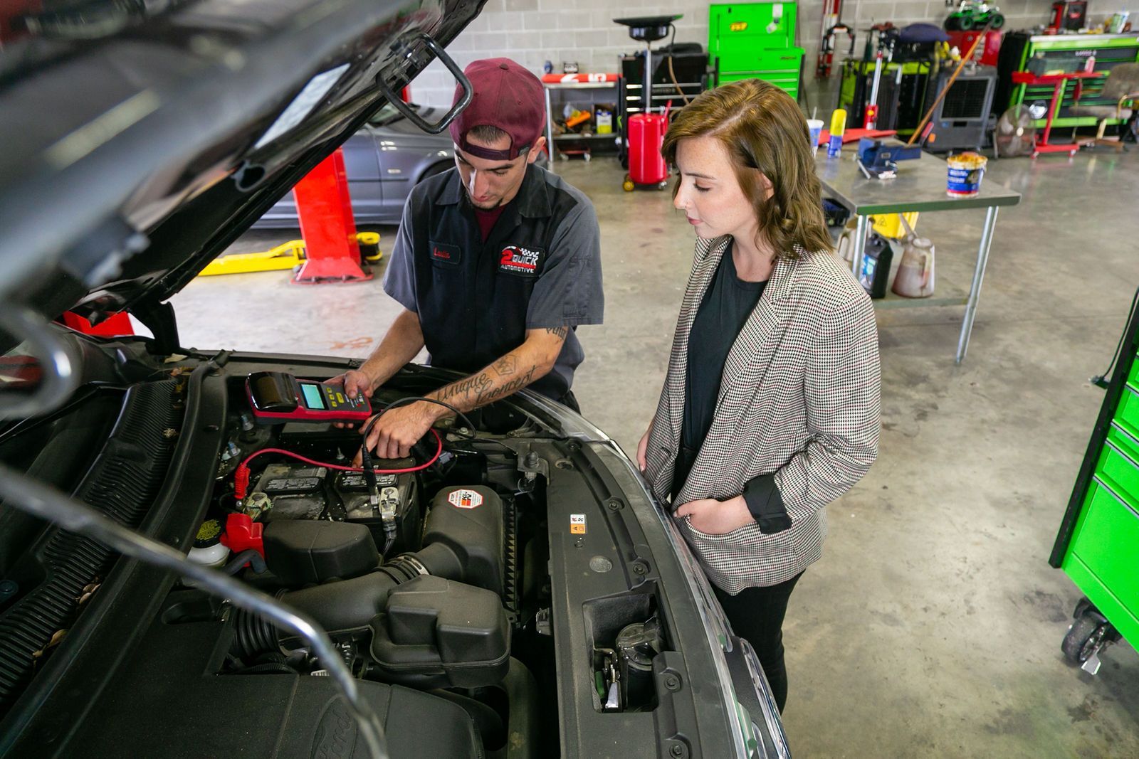 A mechanic tests a car battery with a diagnostic tool while explaining the procedure to a customer in an auto shop. | 2 QuickAuto, Car & Tire Repair | Copperfield