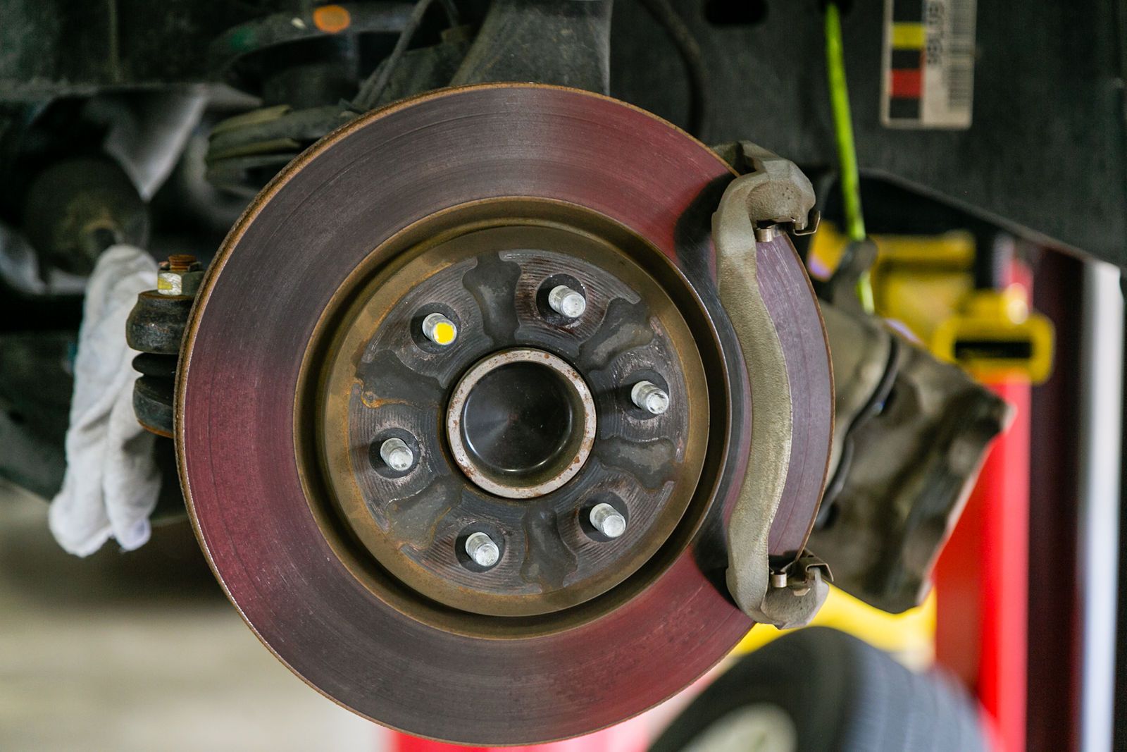 An automobile brake rotor with a rusted, reddish-brown surface, mounted on a vehicle hub assembly in a workshop. | 2 QuickAuto, Car & Tire Repair | Copperfield