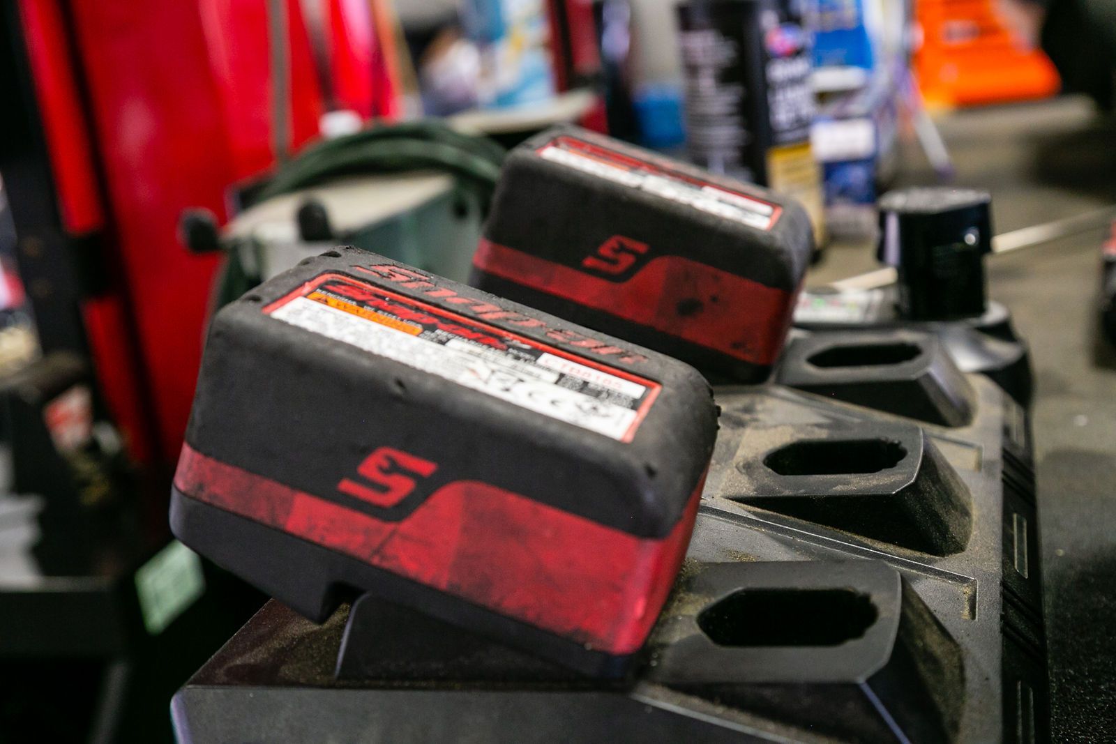 Two red and black Snap-on power tool batteries sitting in a charging station on a workshop workbench. | 2 QuickAuto, Car & Tire Repair | Copperfield