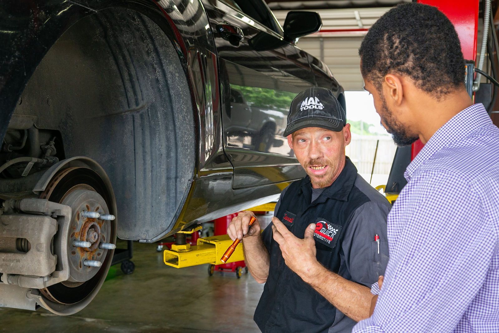 A mechanic in a garage explains car repairs to a customer while pointing at a vehicle's exposed wheel assembly. | 2 QuickAuto, Car & Tire Repair | Copperfield