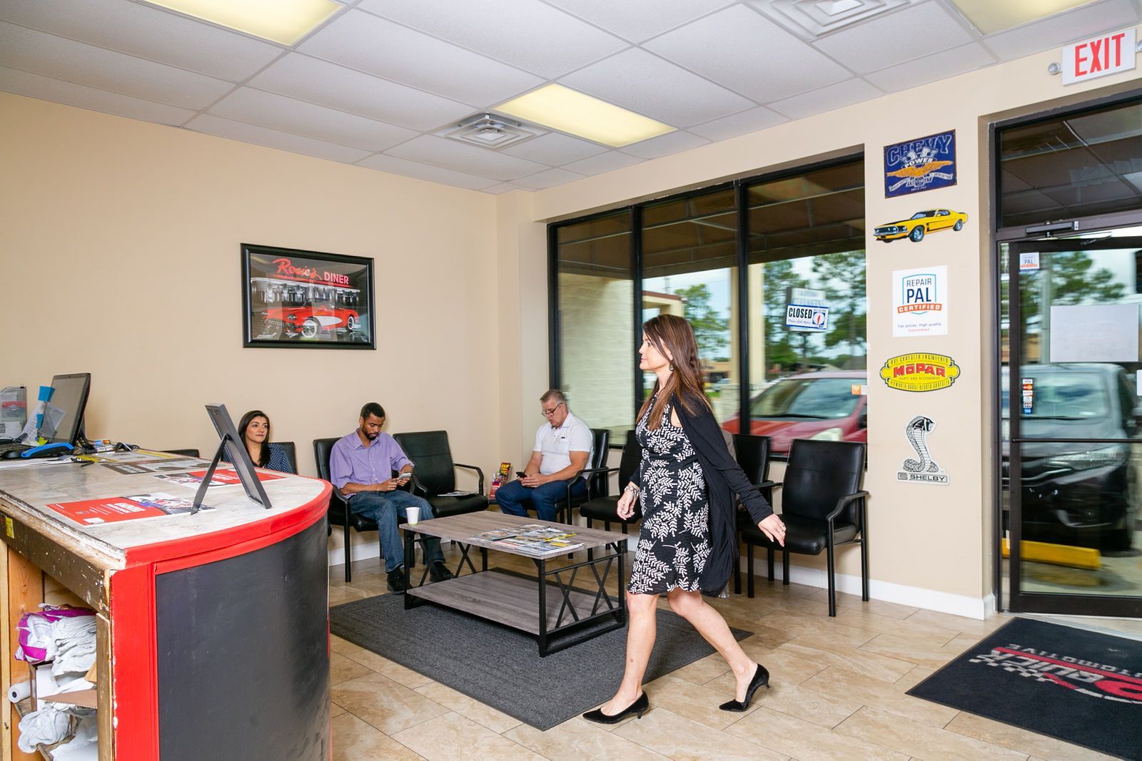 A woman walks through a brightly lit car dealership waiting area with customers seated near a desk and coffee table. | 2 QuickAuto, Car & Tire Repair | Copperfield