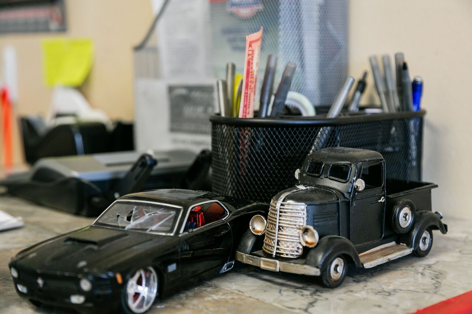 Two black toy vehicles, a modern car and a vintage truck, sit on a desk beside a wire mesh cup filled with pens. | 2 QuickAuto, Car & Tire Repair | Copperfield