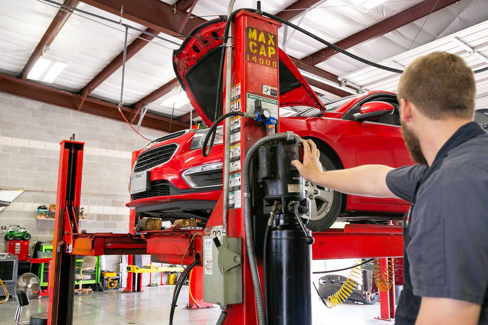 A mechanic in a garage operates a control on a red hydraulic lift holding a red car. | 2 QuickAuto, Car & Tire Repair | Copperfield